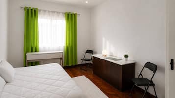 A bedroom with a bed, a desk, a chair, a small table with a lamp, and a potted plant.