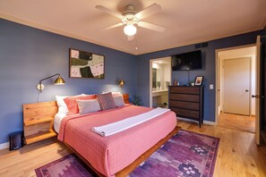 A bedroom with a bed, a ceiling fan, a nightstand, a trash can, a dresser, a mirror, a TV, and a door.