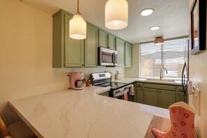 A kitchen with green cabinets, a white marble countertop, a pink coffee maker, and a window with blinds.