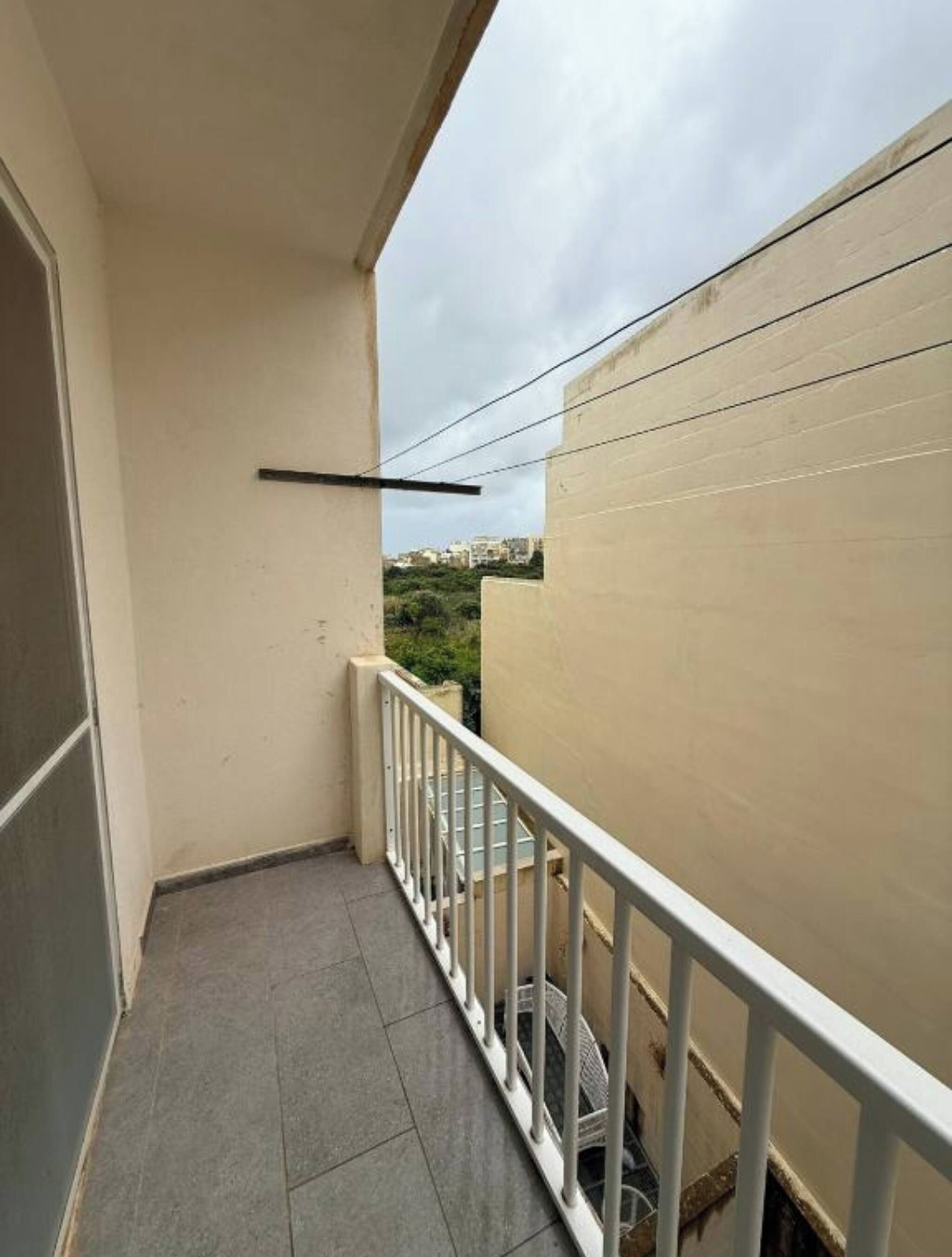 Un balcón estrecho con baranda blanca, piso de baldosas y vista a edificios distantes.