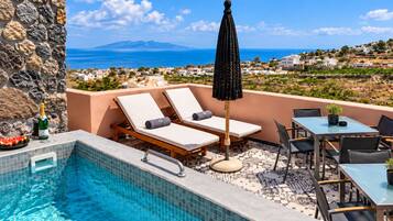 A rooftop pool area with lounge chairs, a table, and a view of the sea and distant hills.