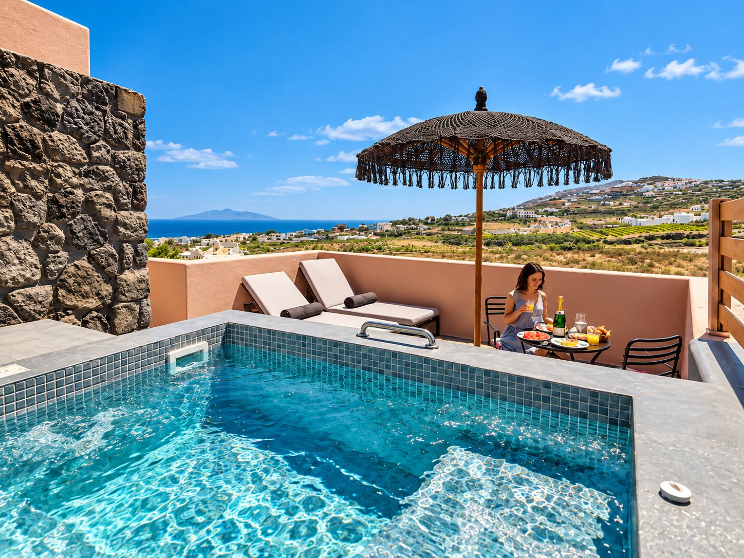 A rooftop pool area with a view of the sea, a stone wall, and a table with food and drinks.