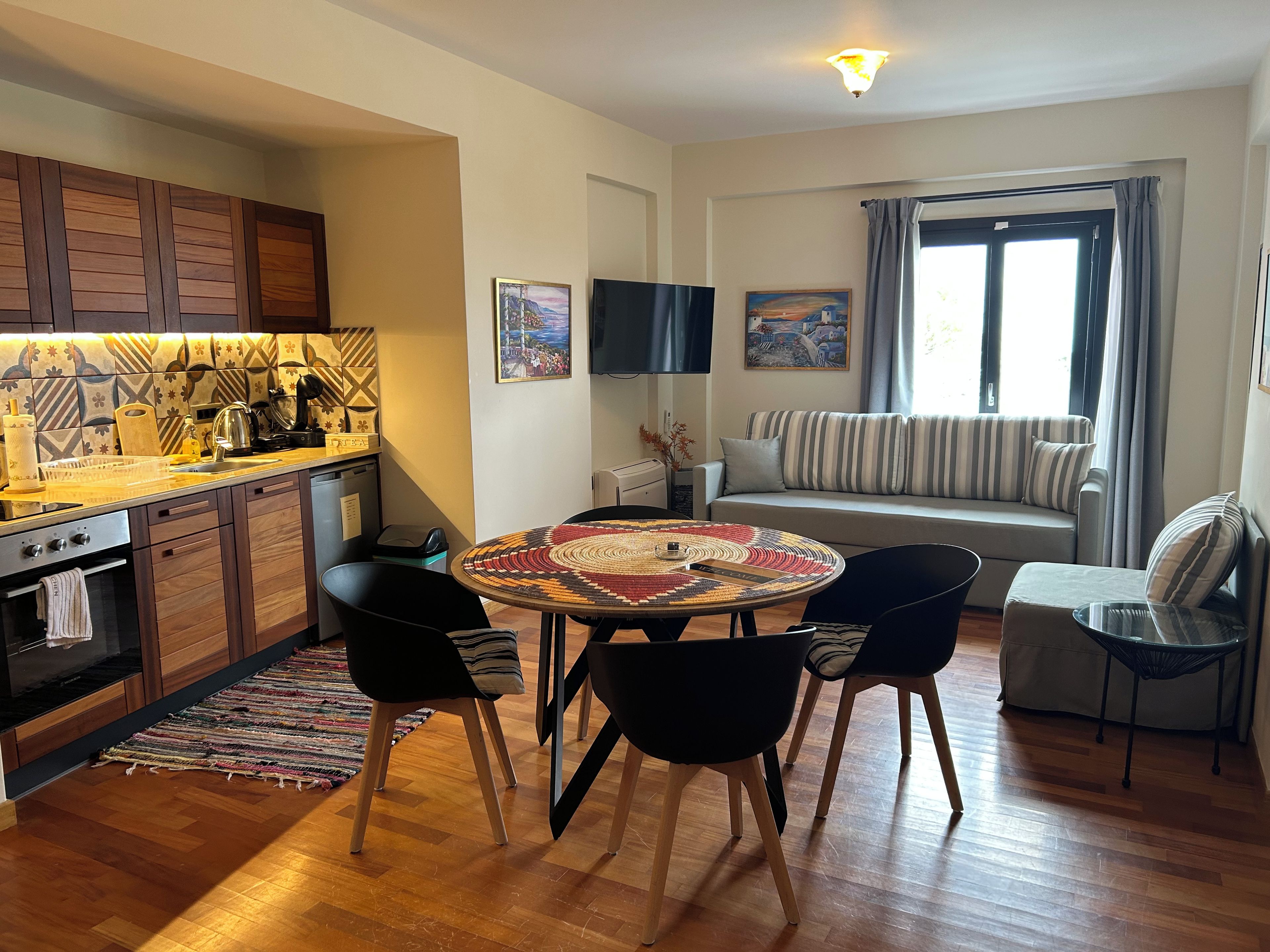 Cozinha moderna com armários de madeira, área de jantar com mesa e cadeiras redondas, sofá e televisão fixada na parede.