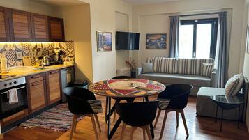 Cozinha moderna com armários de madeira, área de jantar com mesa e cadeiras redondas, sofá e televisão fixada na parede.
