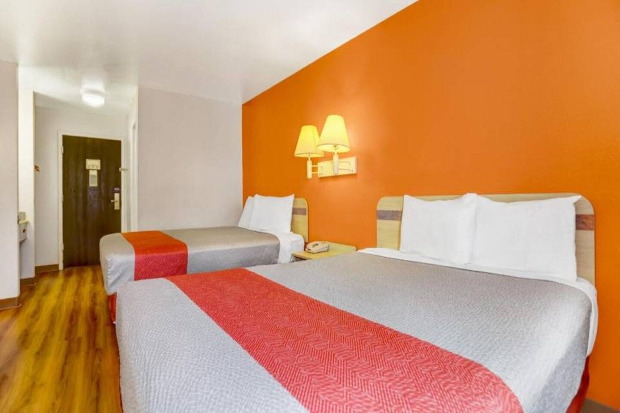A hotel room with two beds, a wooden floor, and a wall-mounted lamp.
