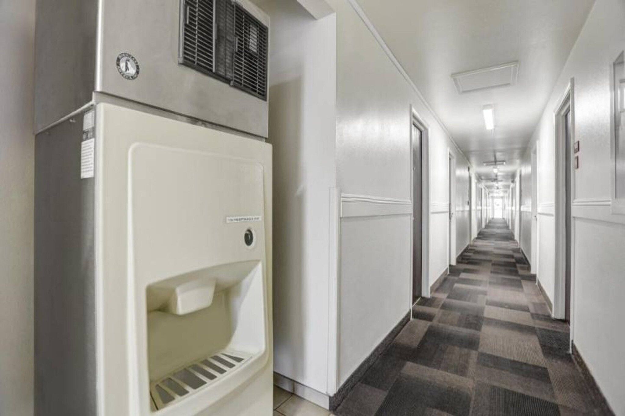 A hotel hallway with a refrigerator on the left, doors on both sides, and a checkered carpet.
