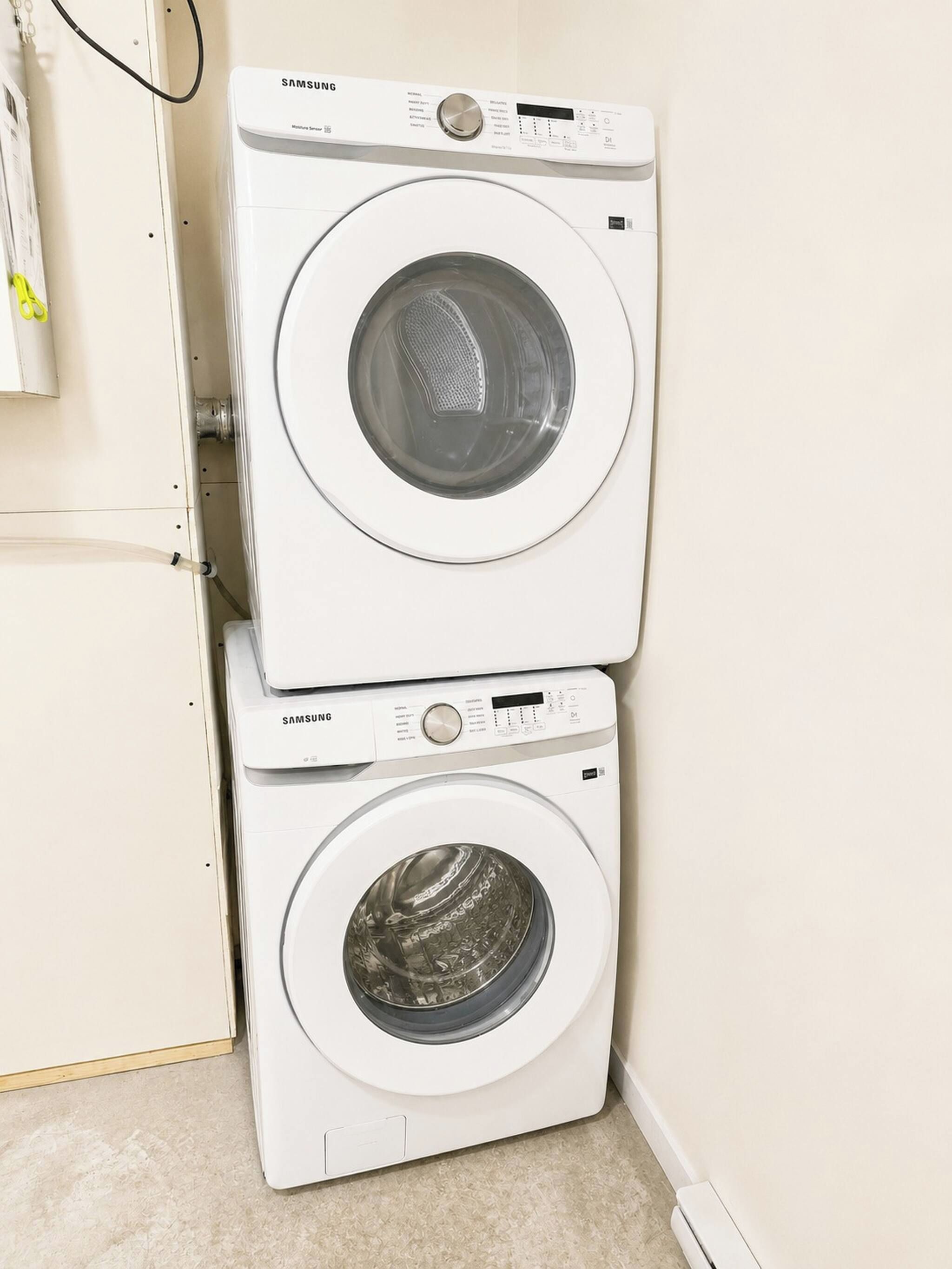 Deux lave-linge Samsung à chargement frontal blancs, placés l’un sur l’autre, dans un coin, avec un sol de couleur beige.