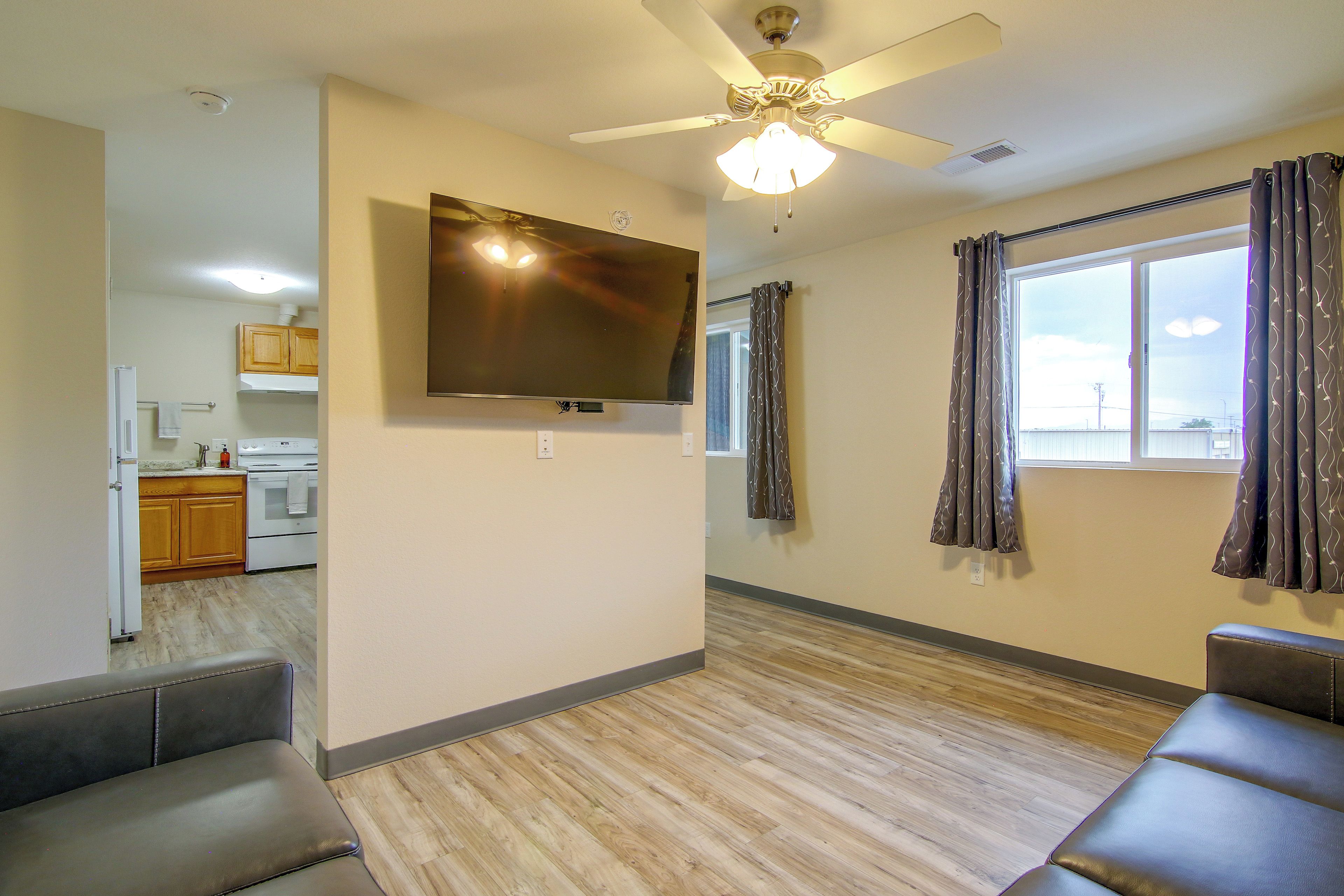 A living room with a flat-screen TV mounted on the wall, a ceiling fan, and a large window with curtains.