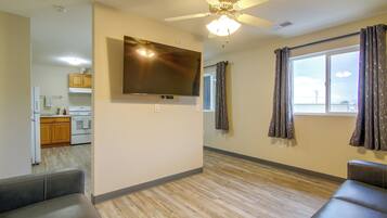 A living room with a flat-screen TV mounted on the wall, a ceiling fan, and a large window with curtains.