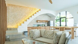 Un intérieur moderne avec un mur en pierre, un canapé agrémenté de coussins à motifs, une table basse en marbre et un coin cuisine doté de meubles en bois et d’une table à manger.