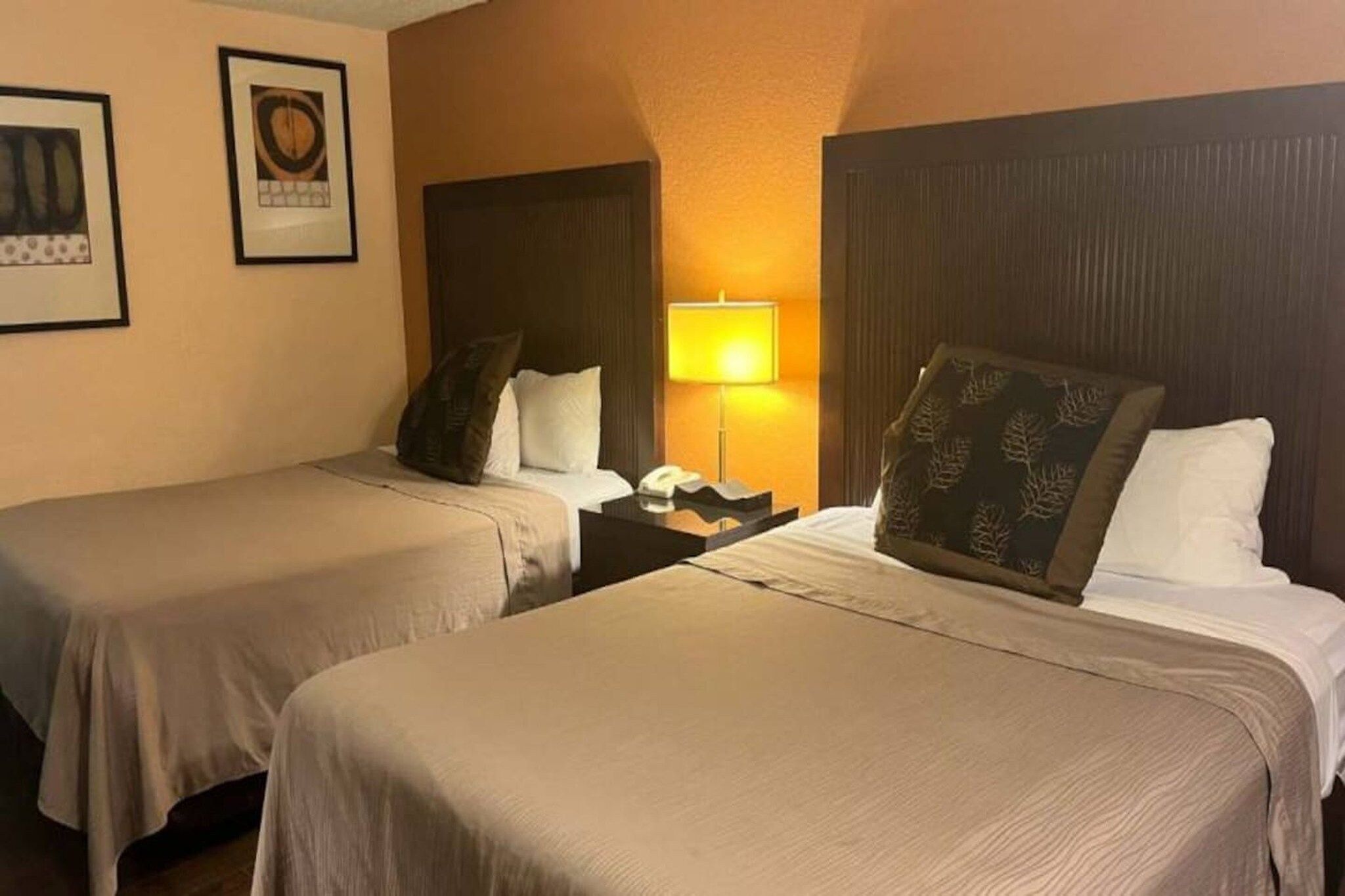 Habitación de hotel con dos camas individuales, una mesita de noche con lámpara y cuadros en la pared.