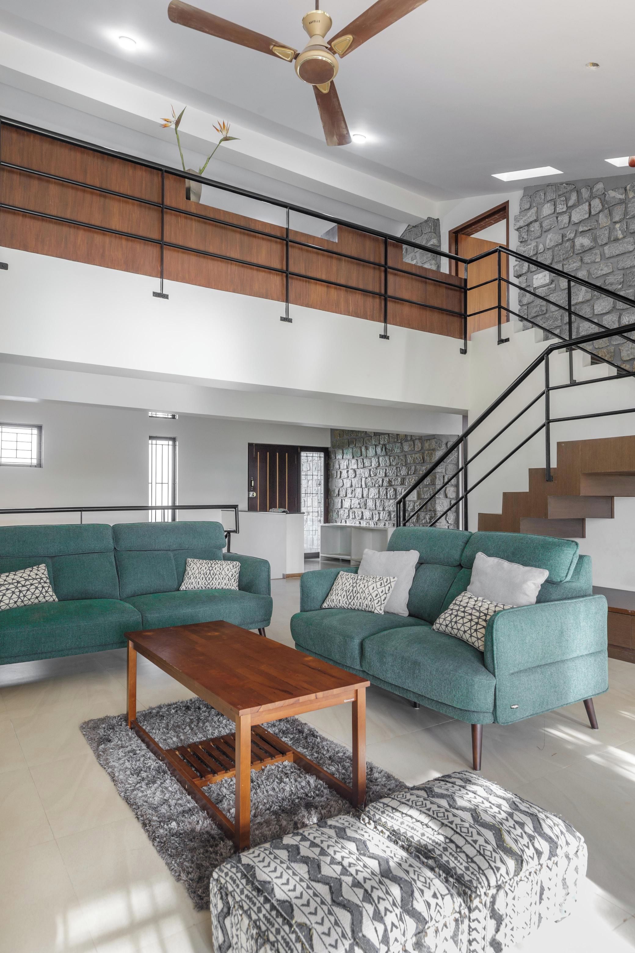 A spacious living area with a high ceiling, a wooden ceiling fan, a staircase with a stone wall, and a green sofa set.