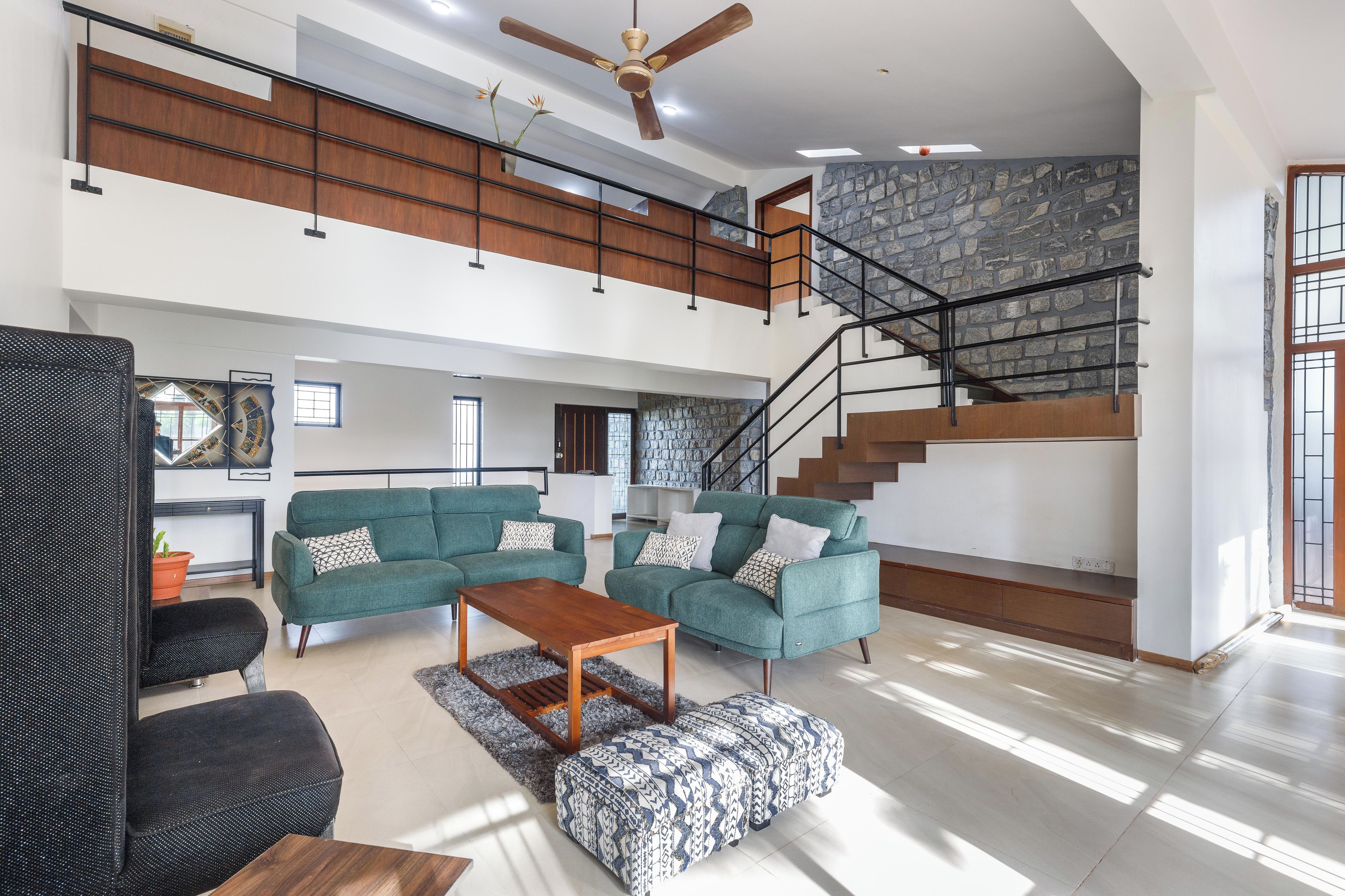 A spacious living area with a green sofa set, a wooden coffee table, and a patterned ottoman. A staircase with a stone wall backdrop leads to an upper level.