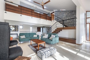 A spacious living area with a green sofa set, a wooden coffee table, and a patterned ottoman. A staircase with a stone wall backdrop leads to an upper level.
