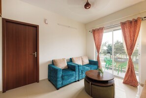 A room with a wooden door, a blue sofa with cushions, a small round table, and a window with patterned curtains. - Lumara by Redolive 2bhk 20min E-city Nr Apollo (Anekal)