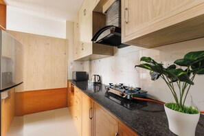 A kitchen with wooden cabinets, a black countertop, a gas stove, a kettle, and a potted plant. - Lumara by Redolive 2bhk 20min E-city Nr Apollo (Anekal)