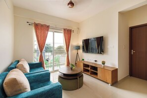 A living room with a blue sofa, a round coffee table, a TV mounted on the wall, and a wooden cabinet. - Lumara by Redolive 2bhk 20min E-city Nr Apollo (Anekal)