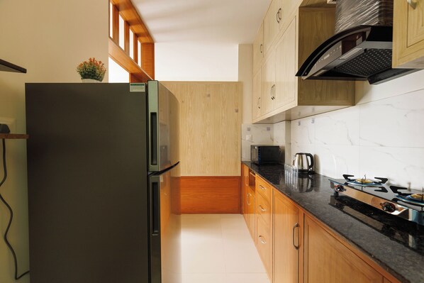 A modern kitchen with a stainless steel refrigerator, wooden cabinets, a black countertop, and a gas stove. - Lumara by Redolive 2bhk 20min E-city Nr Apollo (Anekal)