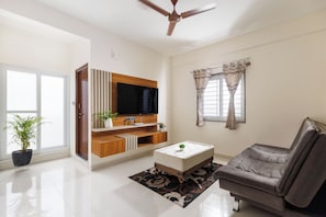 A modern living room with a flat-screen TV, a grey sofa, a coffee table, and a ceiling fan. - Gladia by Redolive 1bhk Nr Manipal Whitefield (Bengaluru)