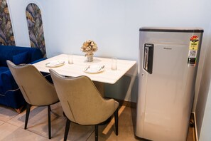 A dining area with a table set for two, two chairs, a refrigerator, and a decorative vase with flowers. - Belmont by Redolive 3bhk 30min DLF Cyber City (Sohna)