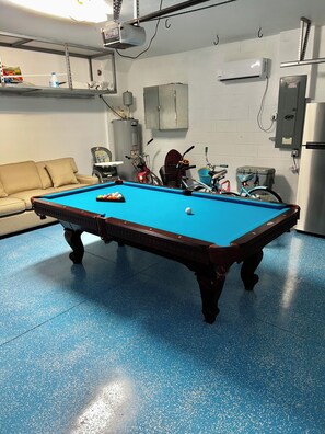 Une table de billard recouverte d’un drap bleu, un canapé et des vélos en arrière-plan.