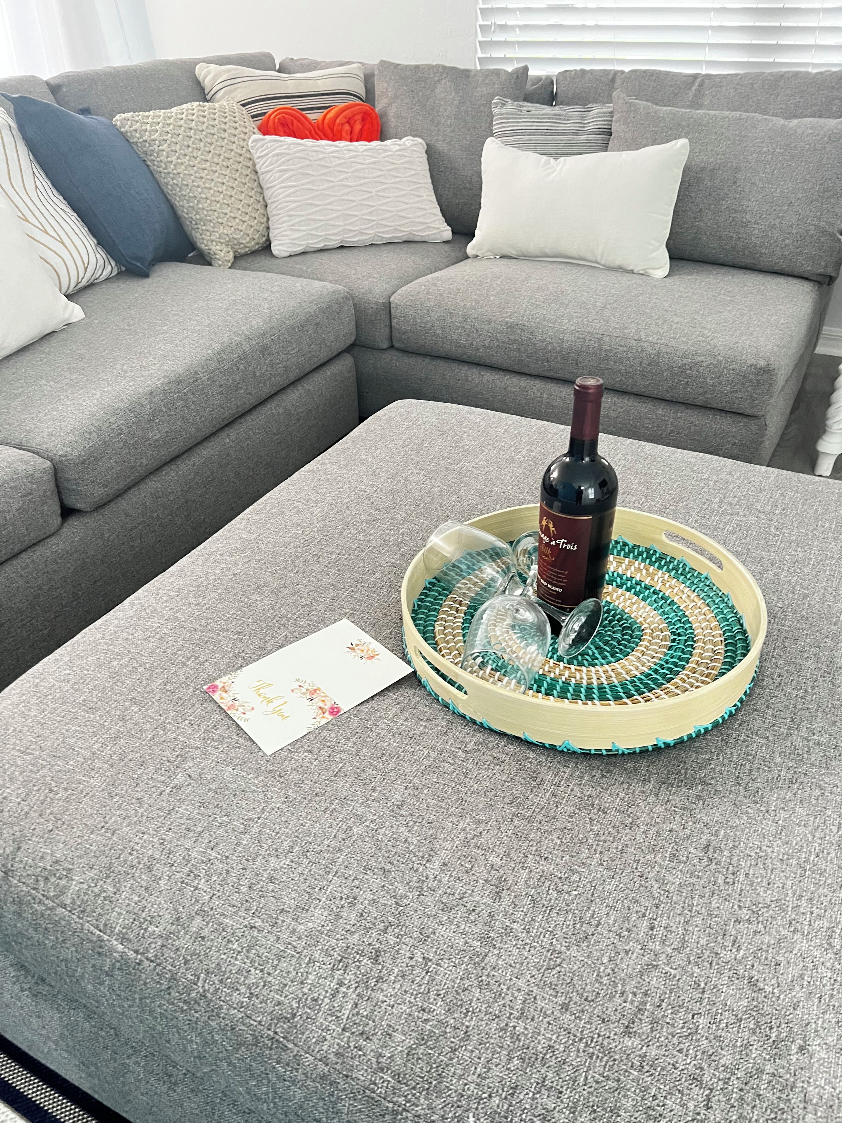 Un canapé gris agrémenté de plusieurs coussins et une bouteille de vin posée sur un plateau rond, accompagnée d’une carte de remerciement.