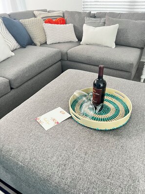 Un canapé gris agrémenté de plusieurs coussins et une bouteille de vin posée sur un plateau rond, accompagnée d’une carte de remerciement.