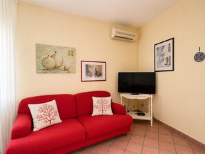 Ein rotes Sofa mit zwei dekorativen Kissen, ein Flachbildfernseher auf einem weißen Standfuß und zwei gerahmte Kunstwerke an der Wand.