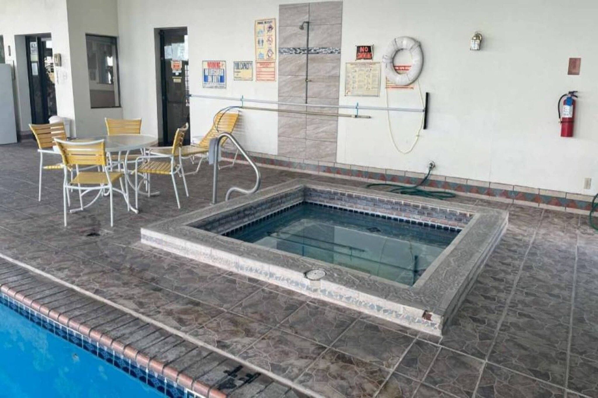 Una piscina con bordeado de baldosas, zona de estar con sillas y señales de seguridad en las paredes.