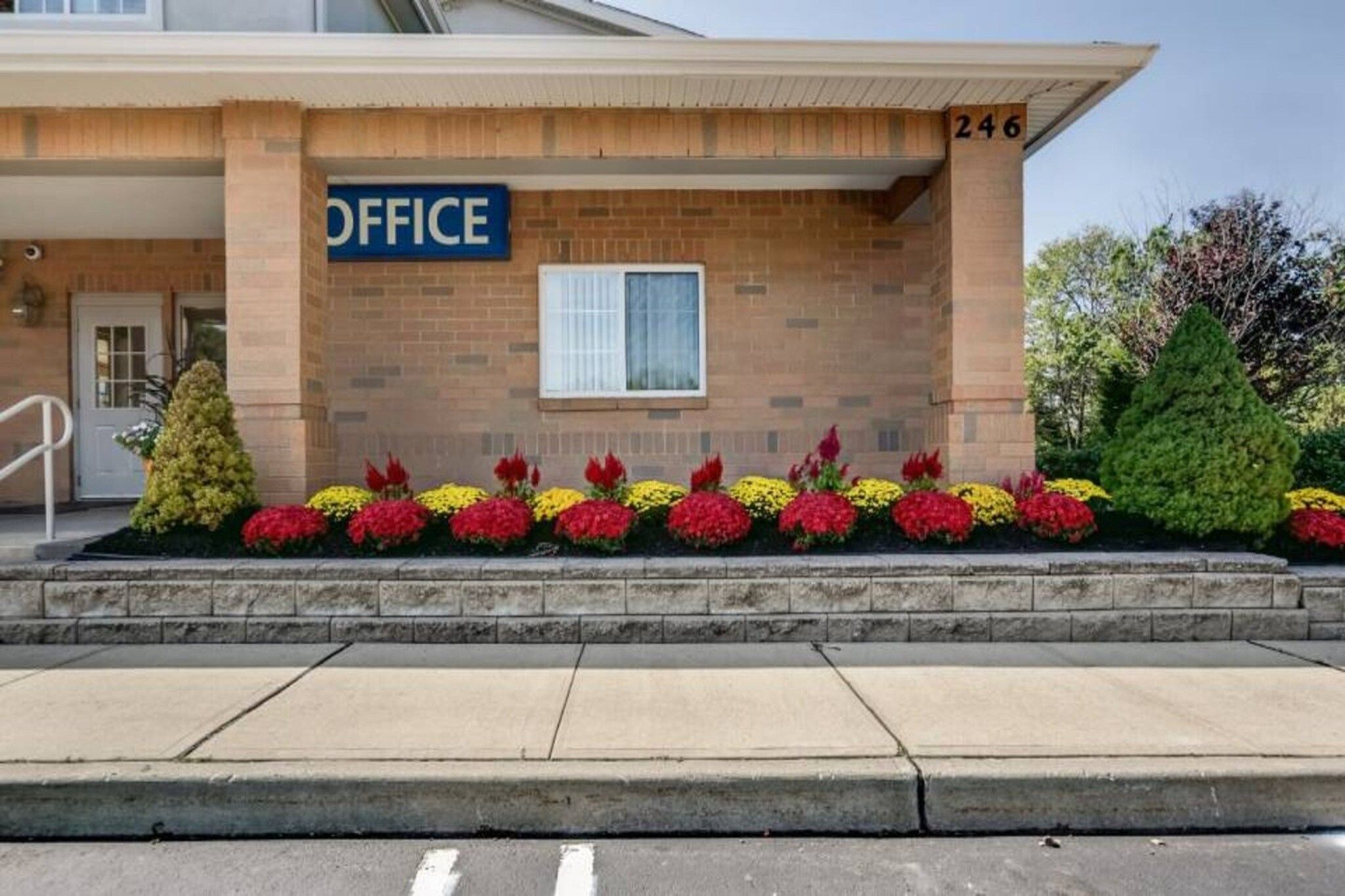 A building with a sign reading 'OFFICE' above the entrance, a brick facade, and a row of colorful flowers in front.