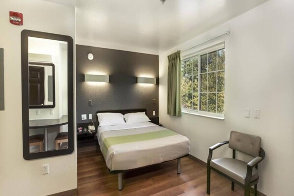 Una camera d'albergo con un letto, una sedia, una finestra con tende, uno specchio e un lampadario a parete.