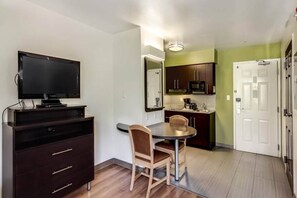 Un espacio habitable compacto con una televisión, una mesa de comedor con sillas, una zona de cocina con armarios y electrodomésticos, y una puerta.