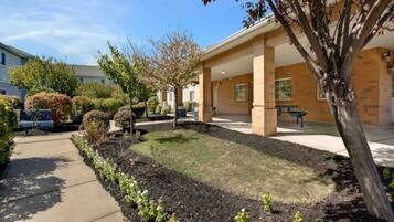 A building with a covered patio area, a sidewalk, and a landscaped garden with shrubs and a tree.