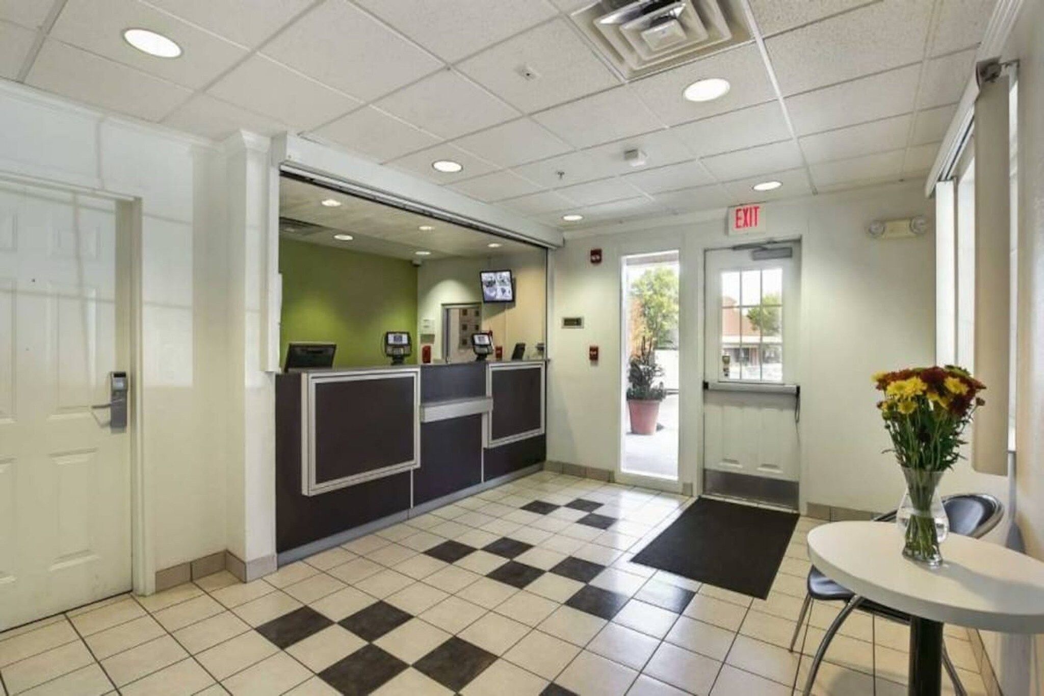 A reception area with a checkered floor, a reception desk, an exit sign, and a small table with a vase of flowers.