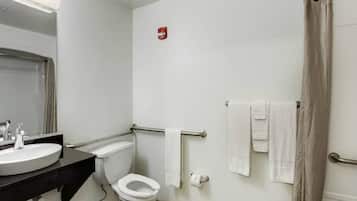 A bathroom with a toilet, sink, and shower. There is a towel rack with towels and a fire alarm on the wall.
