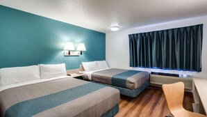 Ett hotellrum med två sängar, ett skrivbord, en stol, ett fönster med gardiner och en vägglampa.
