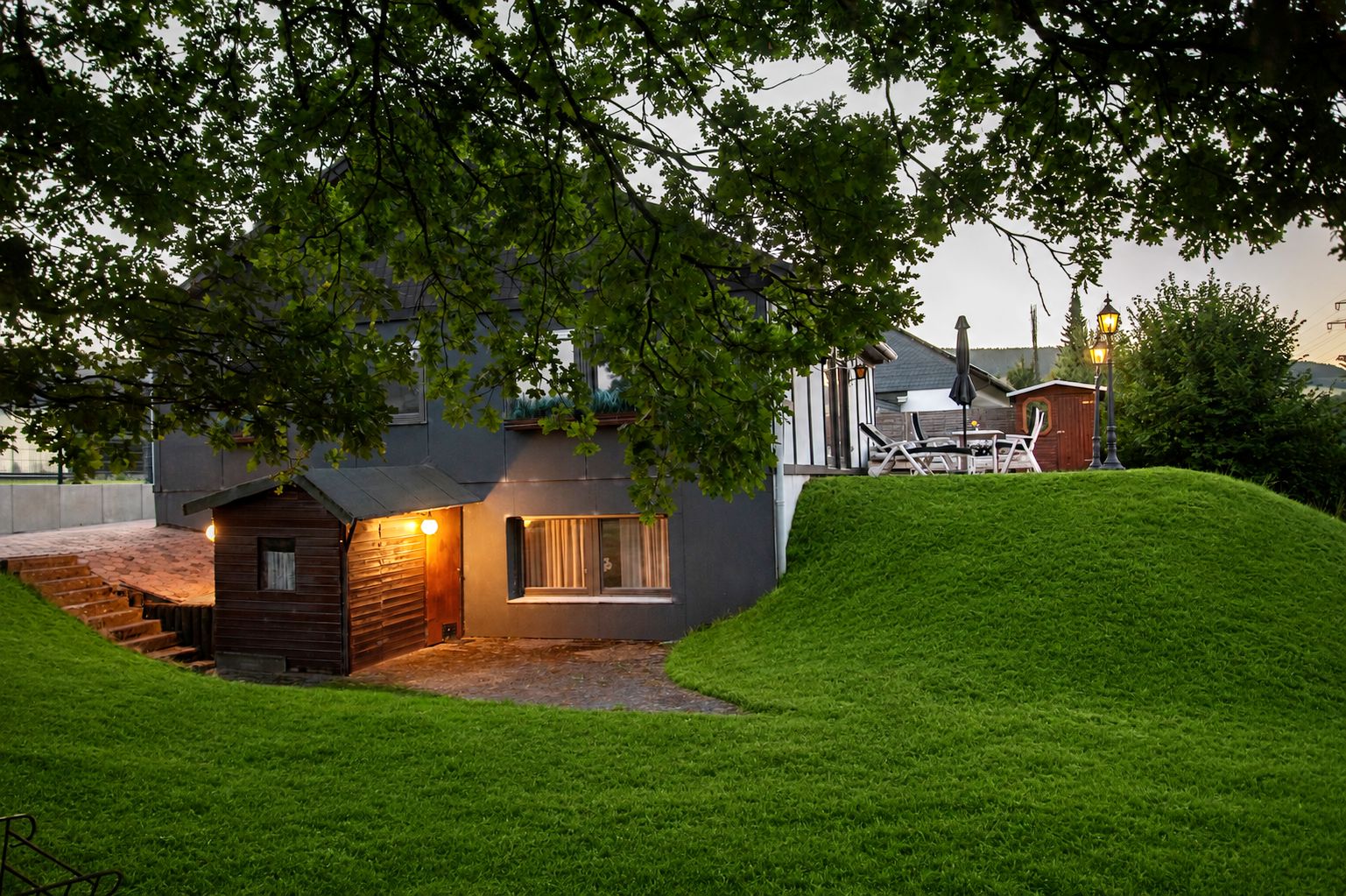 A small wooden cabin with a staircase leading up to the entrance, surrounded by lush green grass and trees.