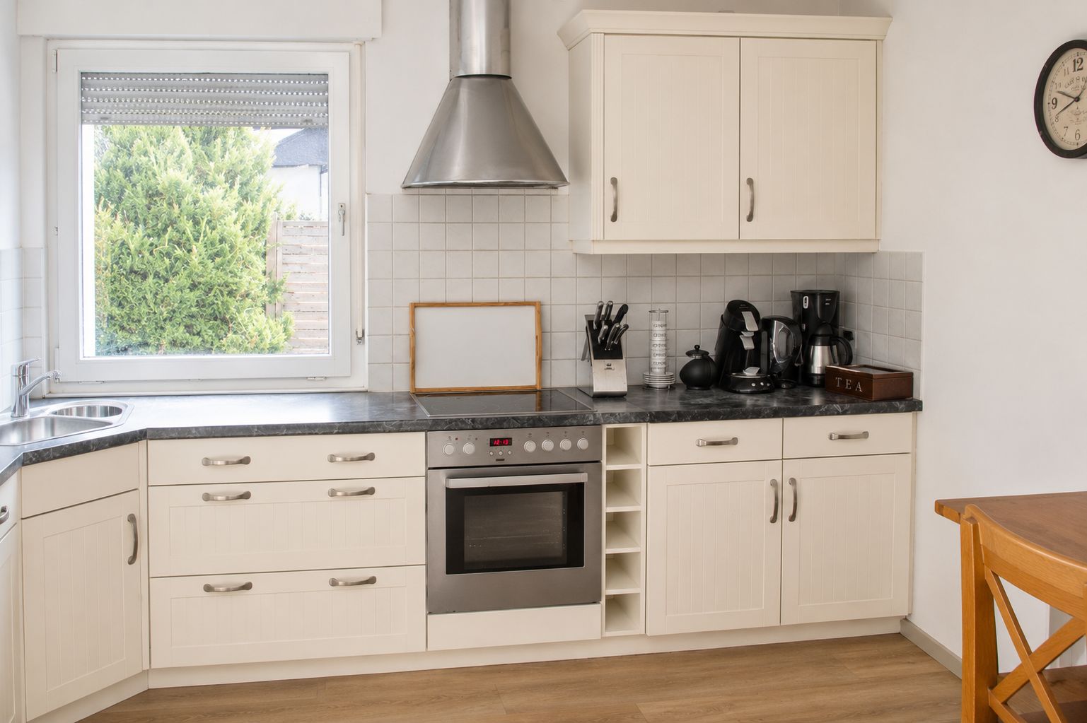 A kitchen with white cabinets, a stainless steel range hood, a built-in oven, a coffee maker, and a window with blinds.