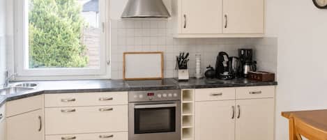 Een keuken met witte kastjes, een roestvrijstalen afzuigkap, een inbouwoven, een koffiezetapparaat en een raam met jaloezieën.