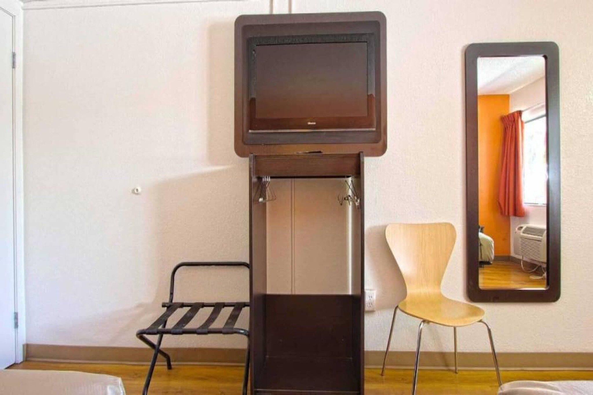 Habitación pequeña con televisión montada en un mueble, una silla y un espejo que refleja una habitación con cama y cortinas.