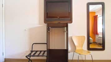 Habitación pequeña con televisión montada en un mueble, una silla y un espejo que refleja una habitación con cama y cortinas.