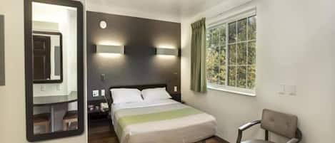 A hotel room with a bed, a chair, a window with curtains, a mirror, and a wall-mounted light fixture.