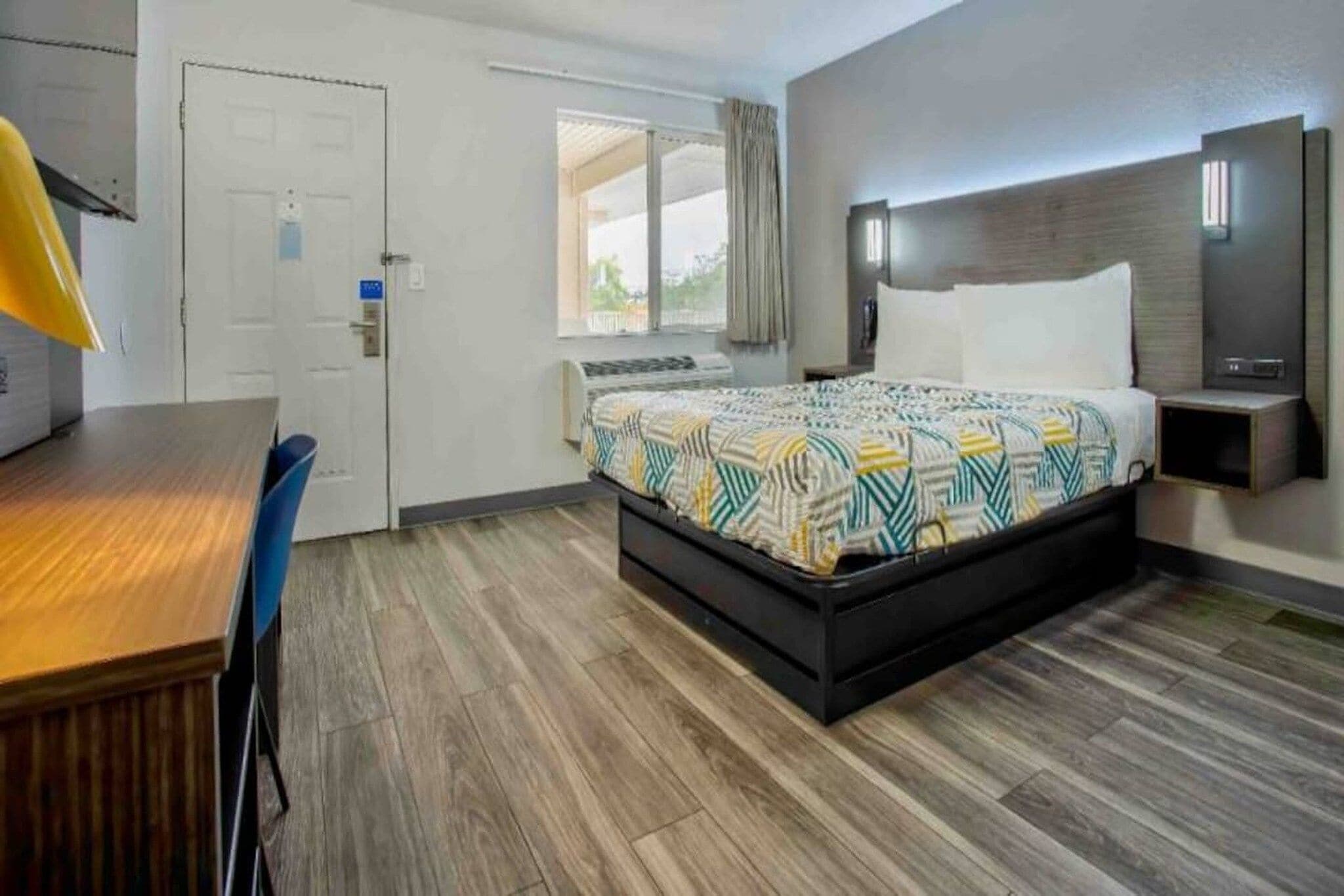 A hotel room with a bed, a desk, a chair, a window with curtains, and a door.