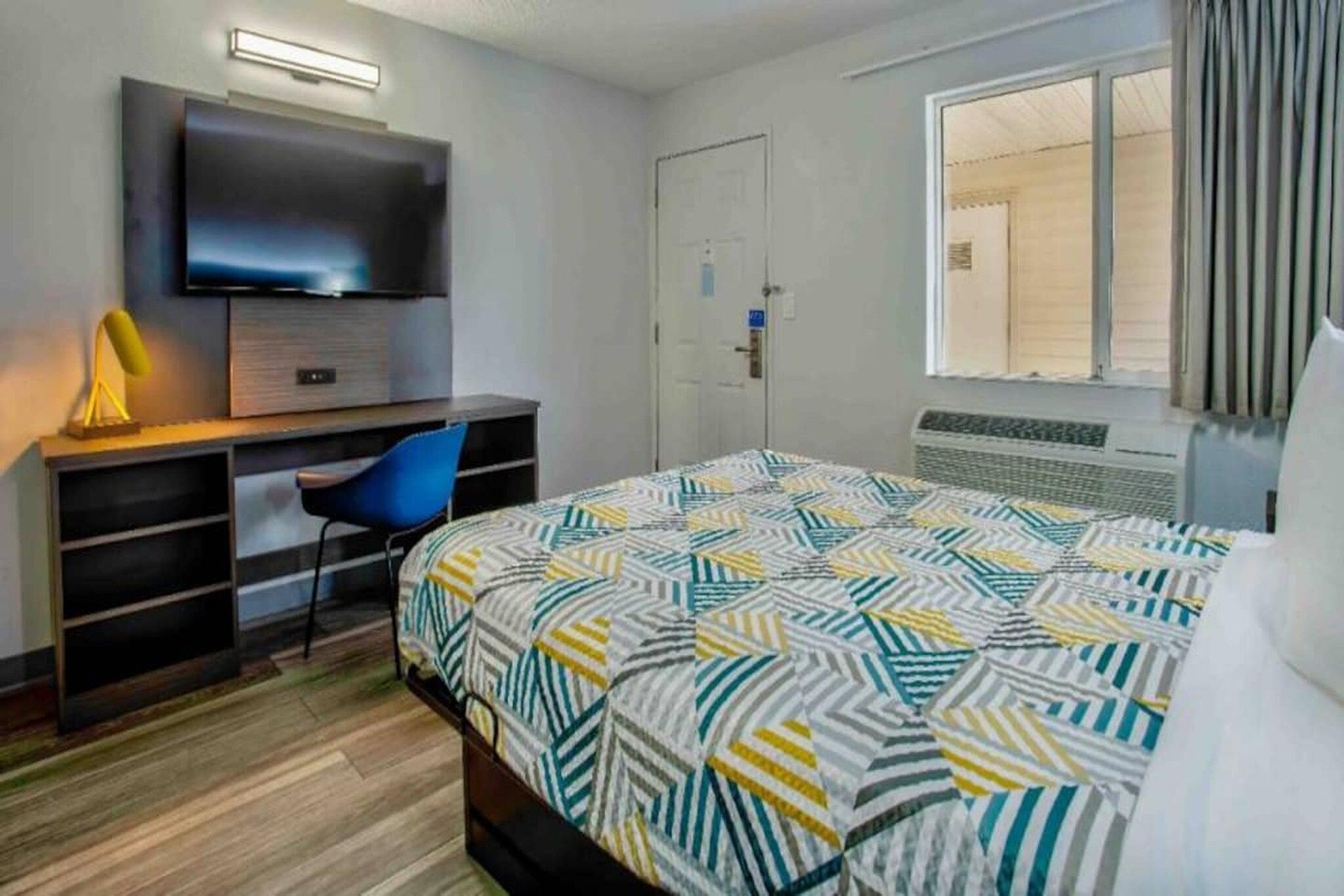 Une chambre d’hôtel avec un lit, un bureau et une chaise, une télévision et une fenêtre avec des rideaux.