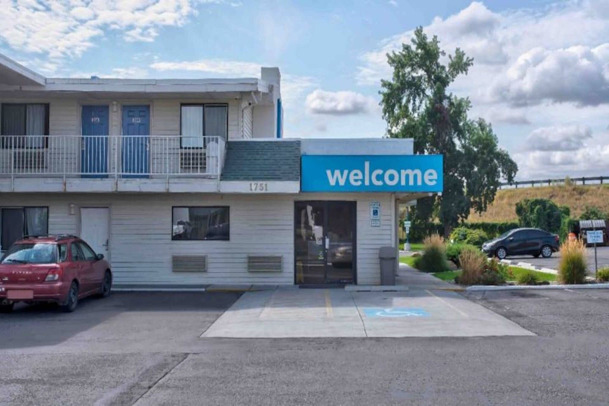 A building with a 'welcome' sign, a parking lot, and a red car parked in front.