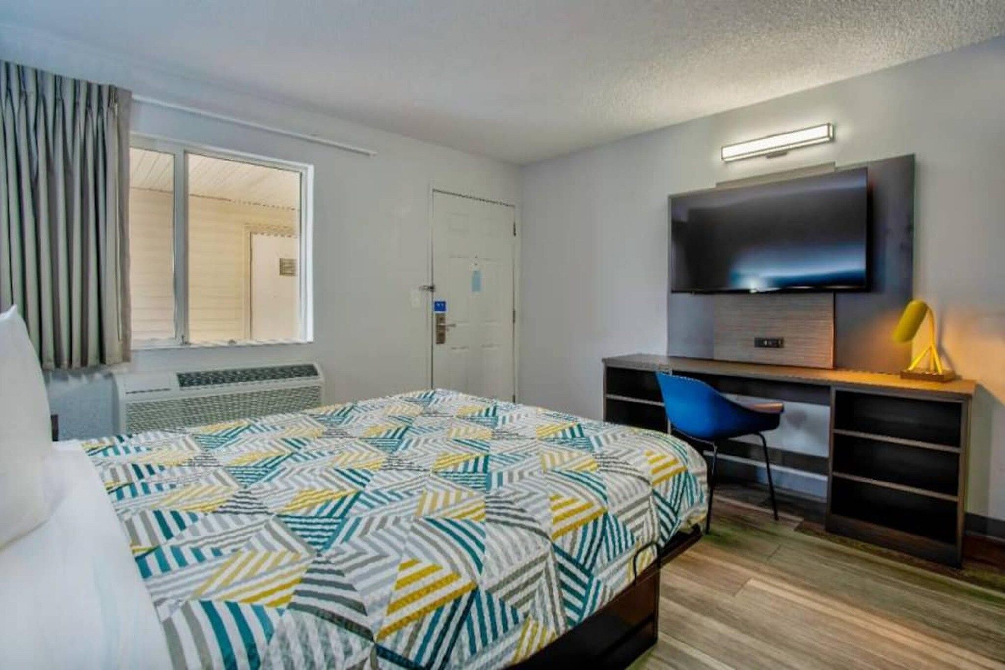 A hotel room with a bed, a desk with a chair, a television, and a window with curtains.