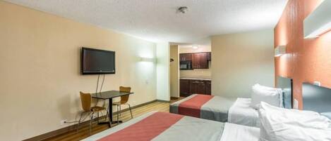 Habitación de hotel con dos camas, un televisor, una pequeña mesa con sillas y una kitchenette al fondo.