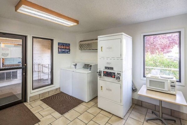 A laundry room with a washing machine, dryer, and microwave. There is a window with a view of trees and a door leading to another room.