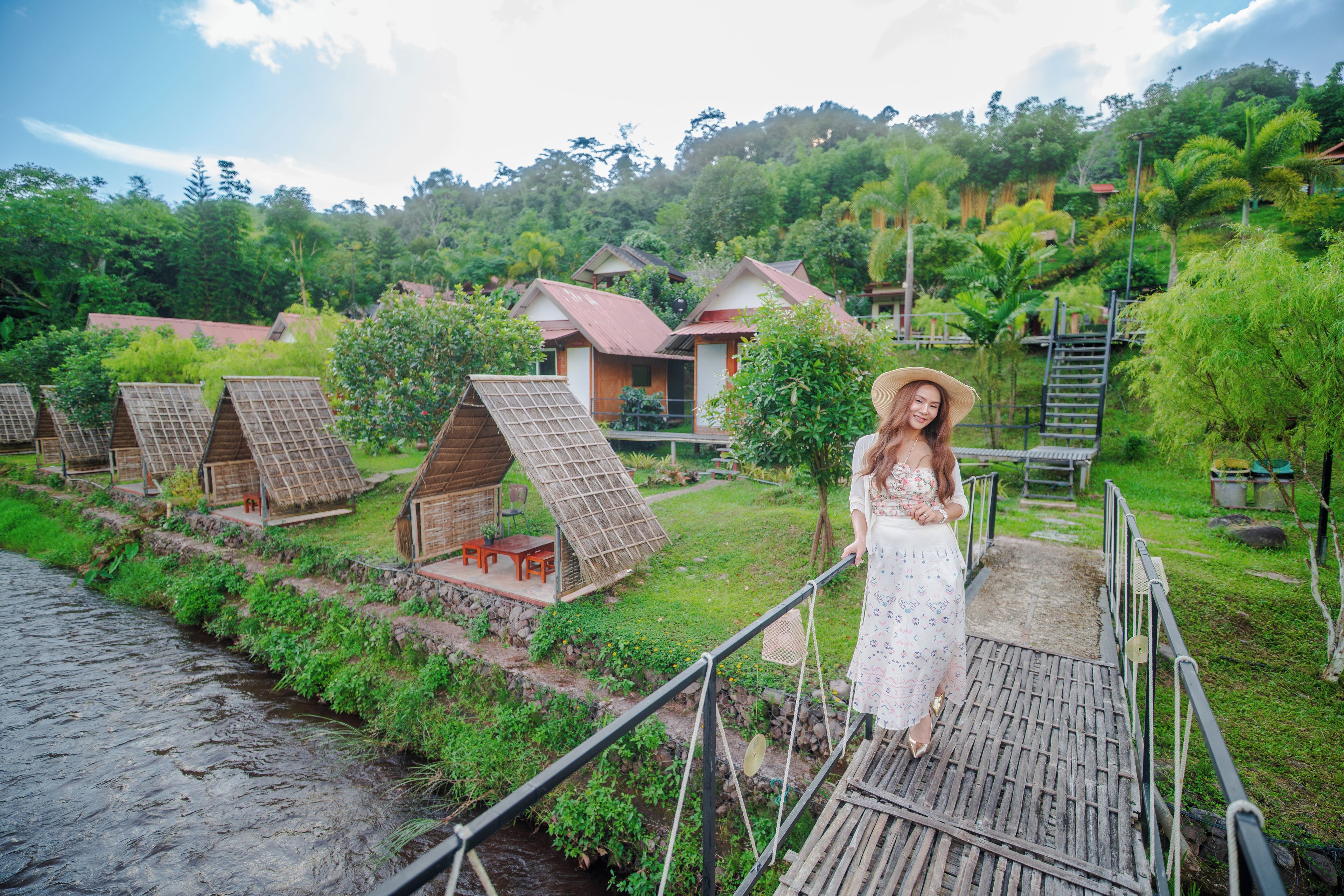 A person standing on a wooden bridge overlooking a series of thatched-roof cottages with a river in the foreground.