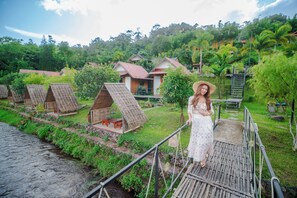 A person standing on a wooden bridge overlooking a series of thatched-roof cottages with a river in the foreground. - Saiwaree Farmstay (Bo Kluea)